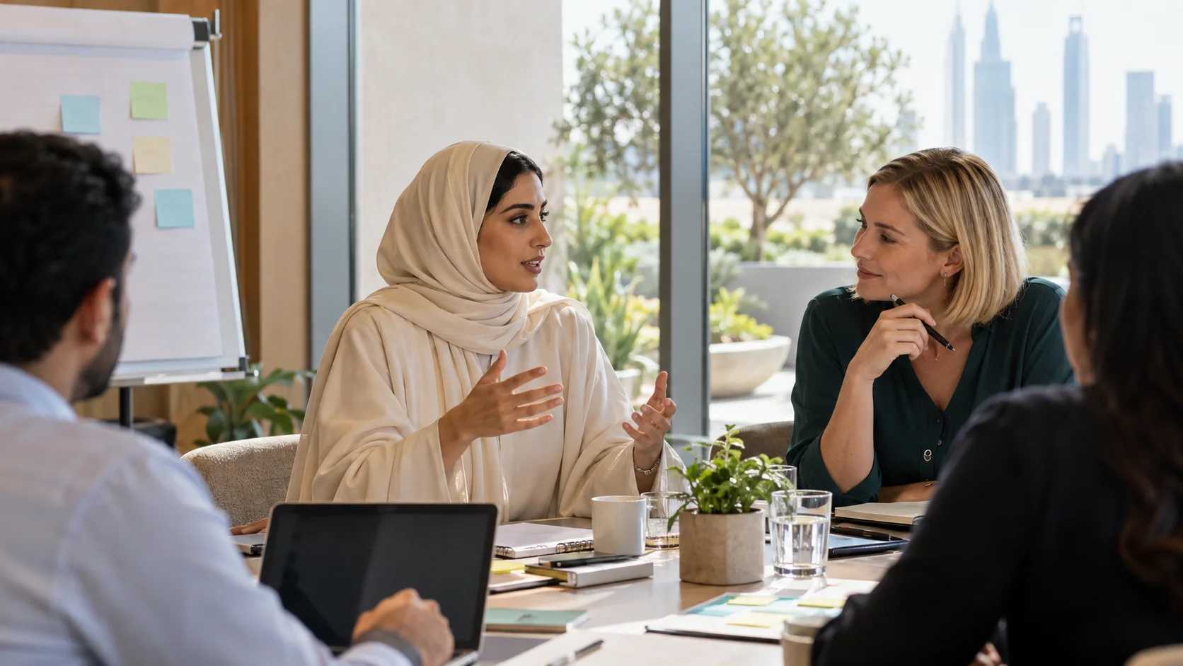 امرأة تتحدث خلال اجتماع حول طاولة عليها حواسيب وأوراق لاصقة، وفي الخارج أفق المدينة.