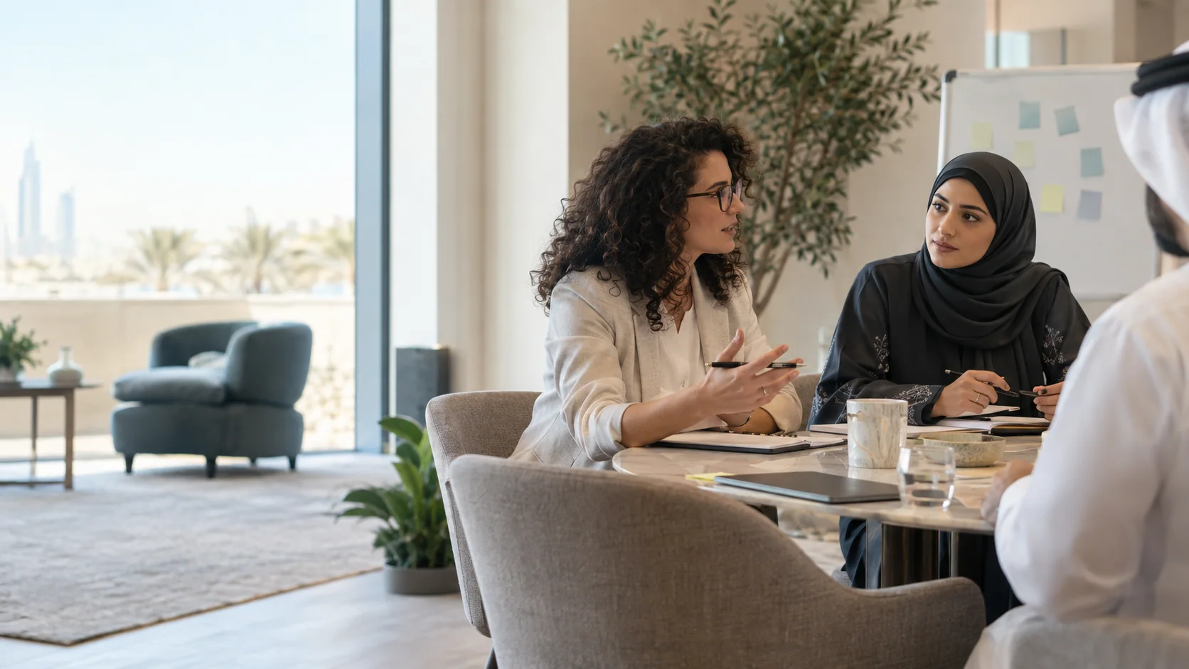 Colleagues talk around a meeting table with notebooks, a whiteboard of sticky notes behind them.