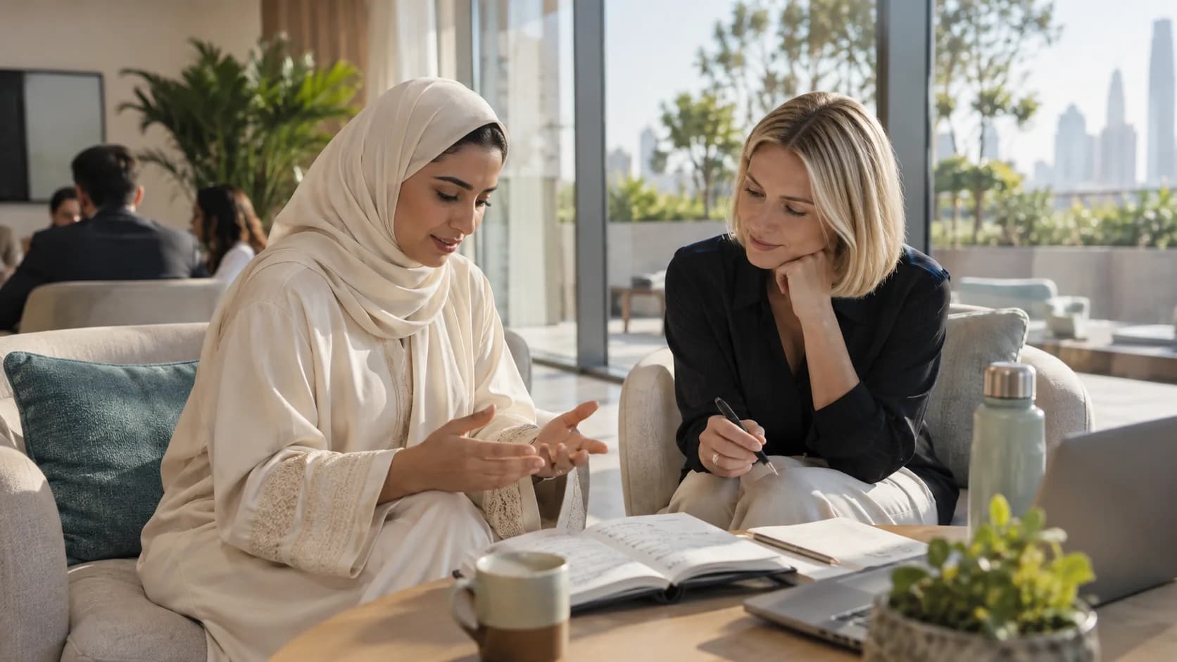 Two women talk through notes in an open notebook in a sunlit lounge overlooking a city skyline.