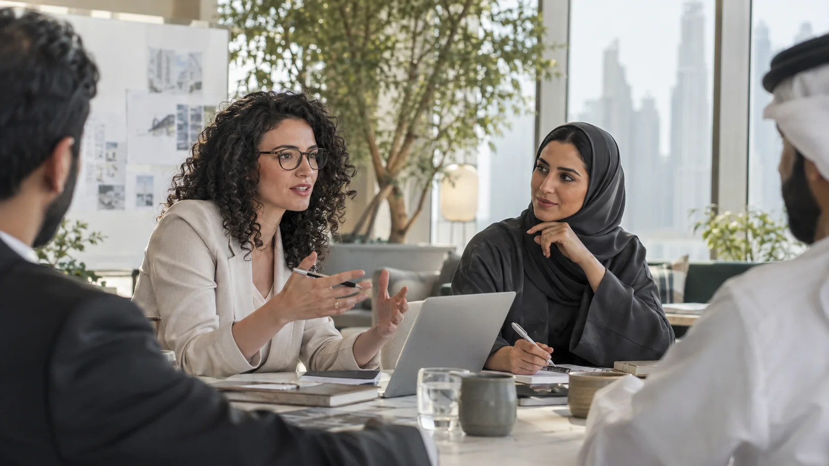 امرأة ترتدي نظارة تتحدث بثقة إلى زملائها حول طاولة اجتماعات، وتظهر من النوافذ أبراج المدينة.