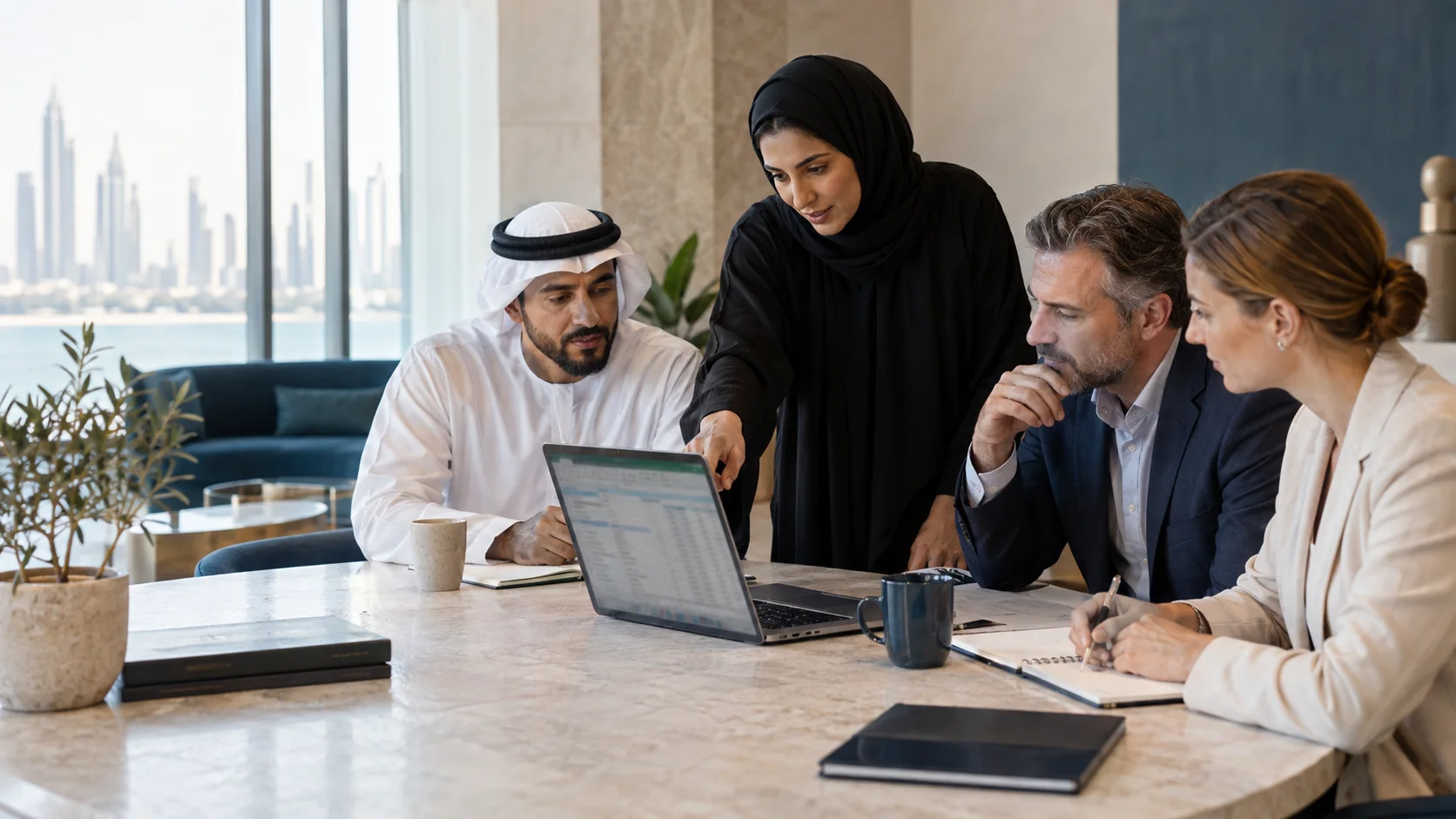 A woman points to a spreadsheet on a laptop as three colleagues at the table follow along.