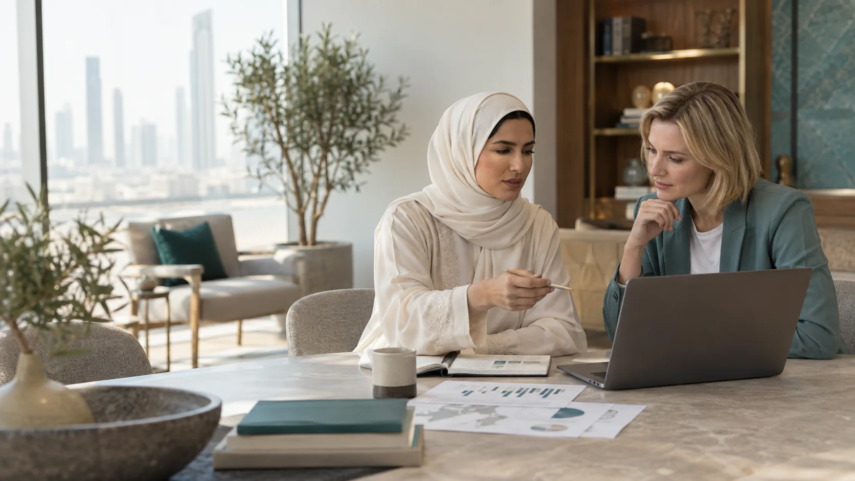 امرأتان تراجعان حاسوبًا محمولًا إلى طاولة عليها رسوم بيانية مطبوعة، وأبراج المدينة تظهر من النافذة.